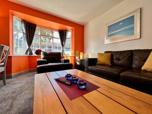 Uma sala de estar com uma mesa de centro de madeira, um sofá de couro marrom, uma janela com cortinas e um quadro na parede.
