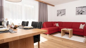A modern living room with a wooden dining table, red sofa, and two black chairs. There are two paintings on the wall and a window with curtains.