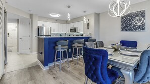 A modern kitchen with stainless steel appliances, a blue accent wall, and a dining area with blue upholstered chairs and a glass table. - Cozy N. Myrtle Beach Condo w/ access to resort amenities. Short Walk to Beach.   (North Myrtle Beach)
