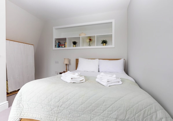 Una cama bien hecha con toallas dobladas, una mesita de noche con una lámpara y una estantería con libros y objetos decorativos.