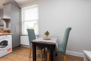 Una cocina compacta con lavadora, una mesa con mantel a rayas y dos sillas tapizadas en verde.
