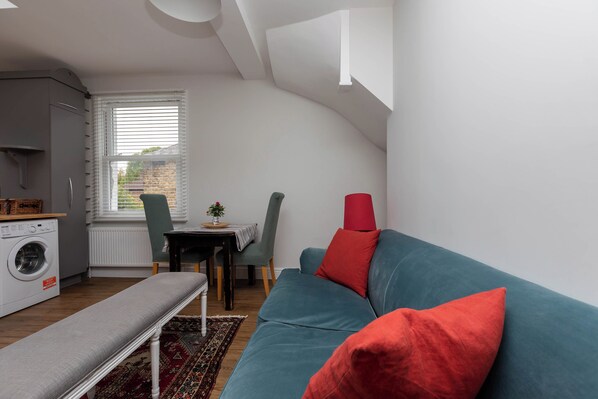 Un espacio habitable compacto con lavadora, zona de comedor con mesa y sillas, sofá con cojines y una banca.
