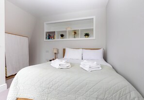 Una cama bien hecha con sábanas blancas y un edredón acolchado, una mesita de noche con lámpara, y un estante montado en la pared con libros y objetos decorativos.