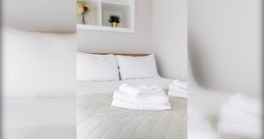 A hotel room with a neatly made bed, white bedding, and a small shelf with flowers.