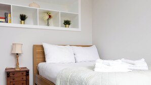 Una cama bien hecha con ropa blanca, una mesita de noche con lámpara y un estante sobre la cama con libros y adornos.