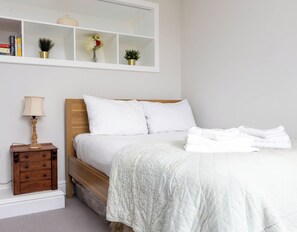 A neatly made bed with white bedding, a bedside table with a lamp, and a shelf above the bed with books and decorative items.