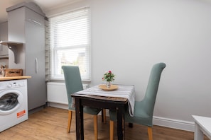 A compact kitchen with a washing machine, a table with a striped tablecloth, and two chairs with a woven texture.