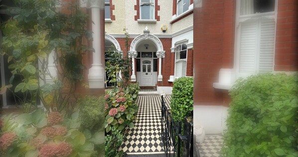 A courtyard with a checkered floor leading to a white door, surrounded by greenery and brick walls.