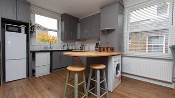 A modern kitchen with grey cabinets, a wooden island with stools, a washing machine, and a window with blinds.