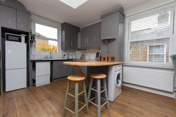 A modern kitchen with grey cabinets, a wooden island with stools, a washing machine, and a window with blinds.