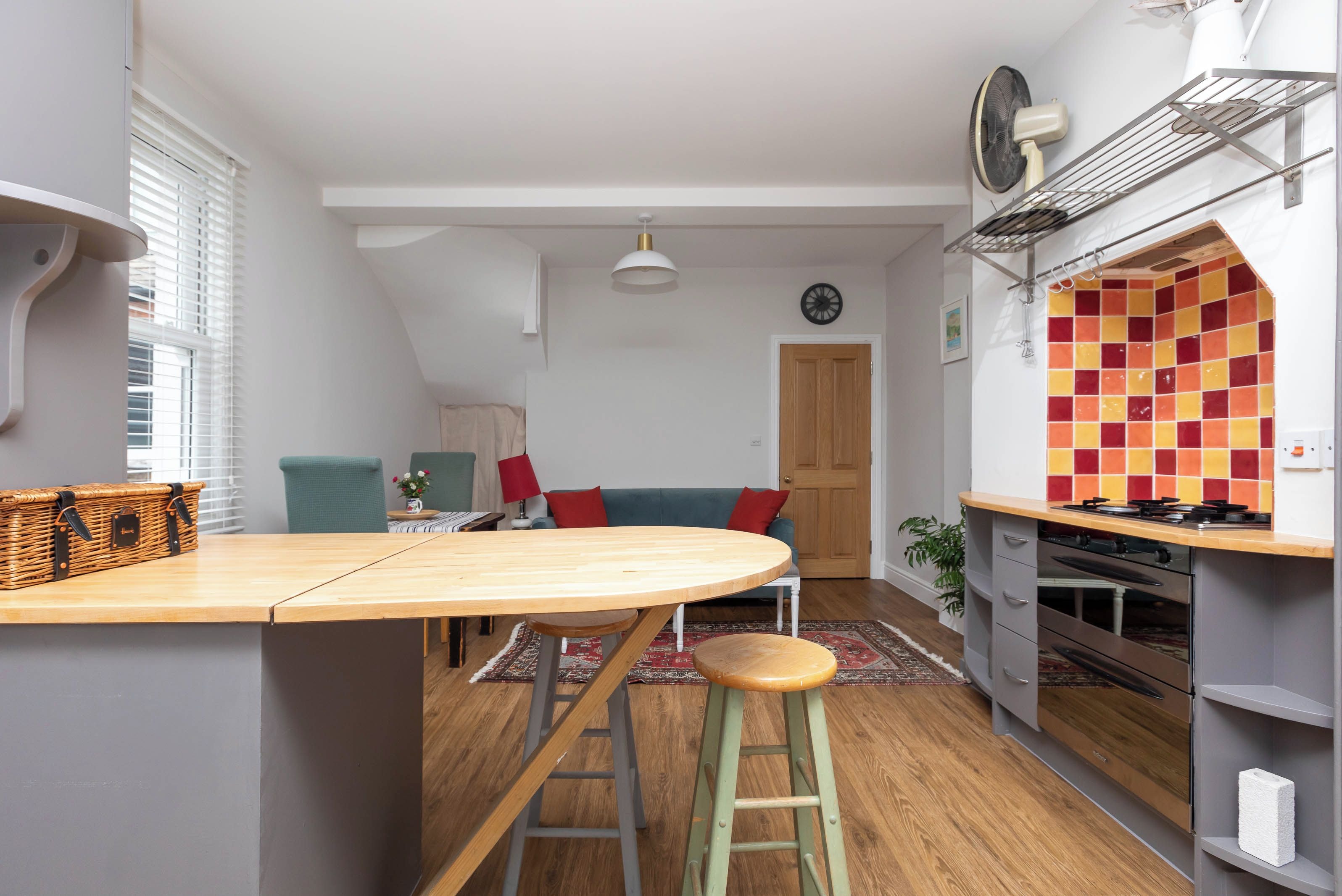 Una cocina con isla de madera, taburetes, un salpicadero de azulejos rojos y amarillos, y vistas al área de estar con un sofá y una puerta.