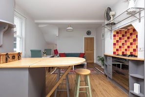 A kitchen with a wooden island, stools, a red and yellow tiled backsplash, and a view to the living area with a sofa and a door.