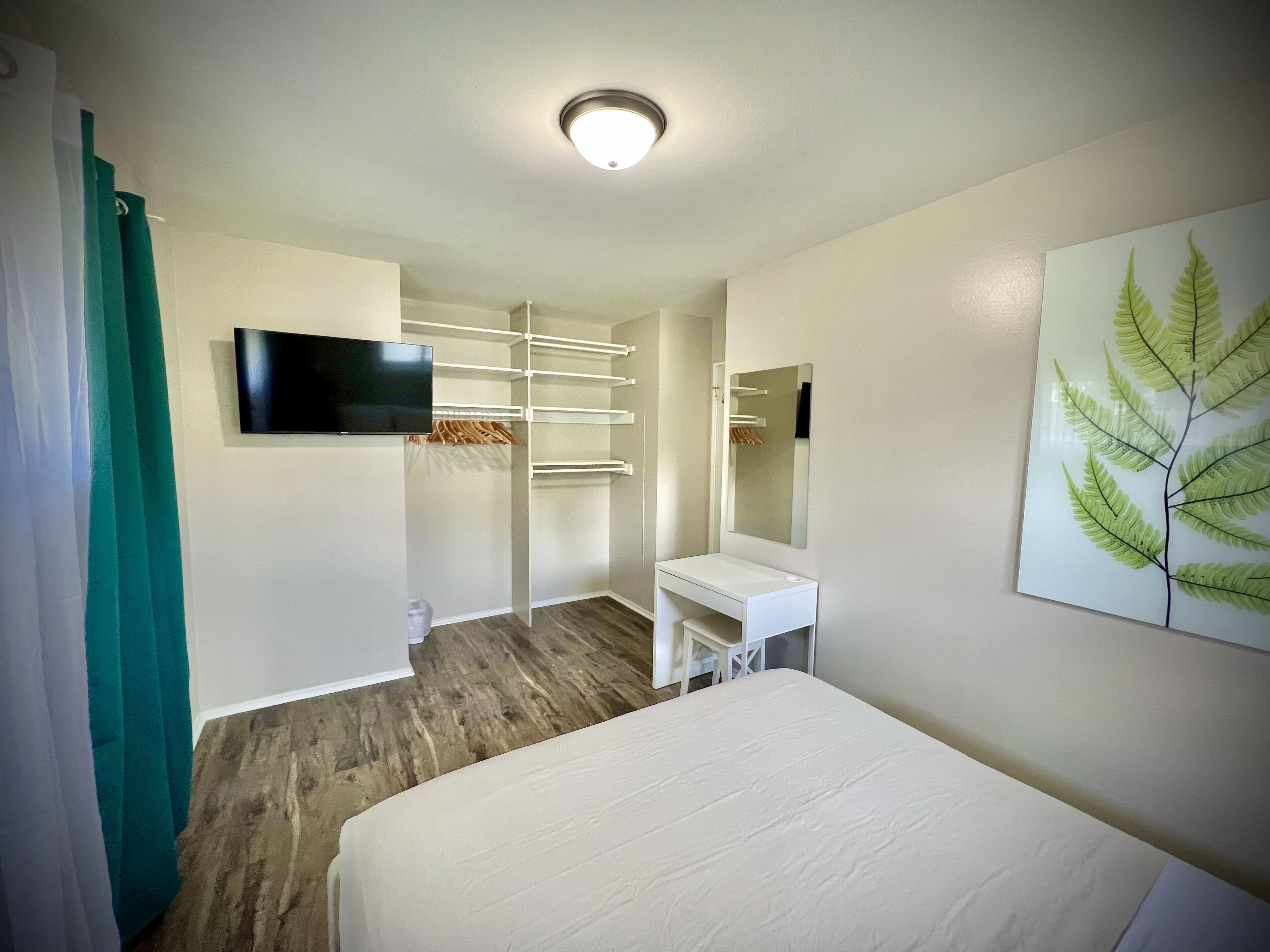 Une chambre d’hôtel avec un lit, une télévision, un bureau blanc, un miroir et une armoire avec des étagères.