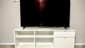 A flat-screen TV on a white TV stand with two shelves and a drawer, against a plain wall with a thermostat on the left.