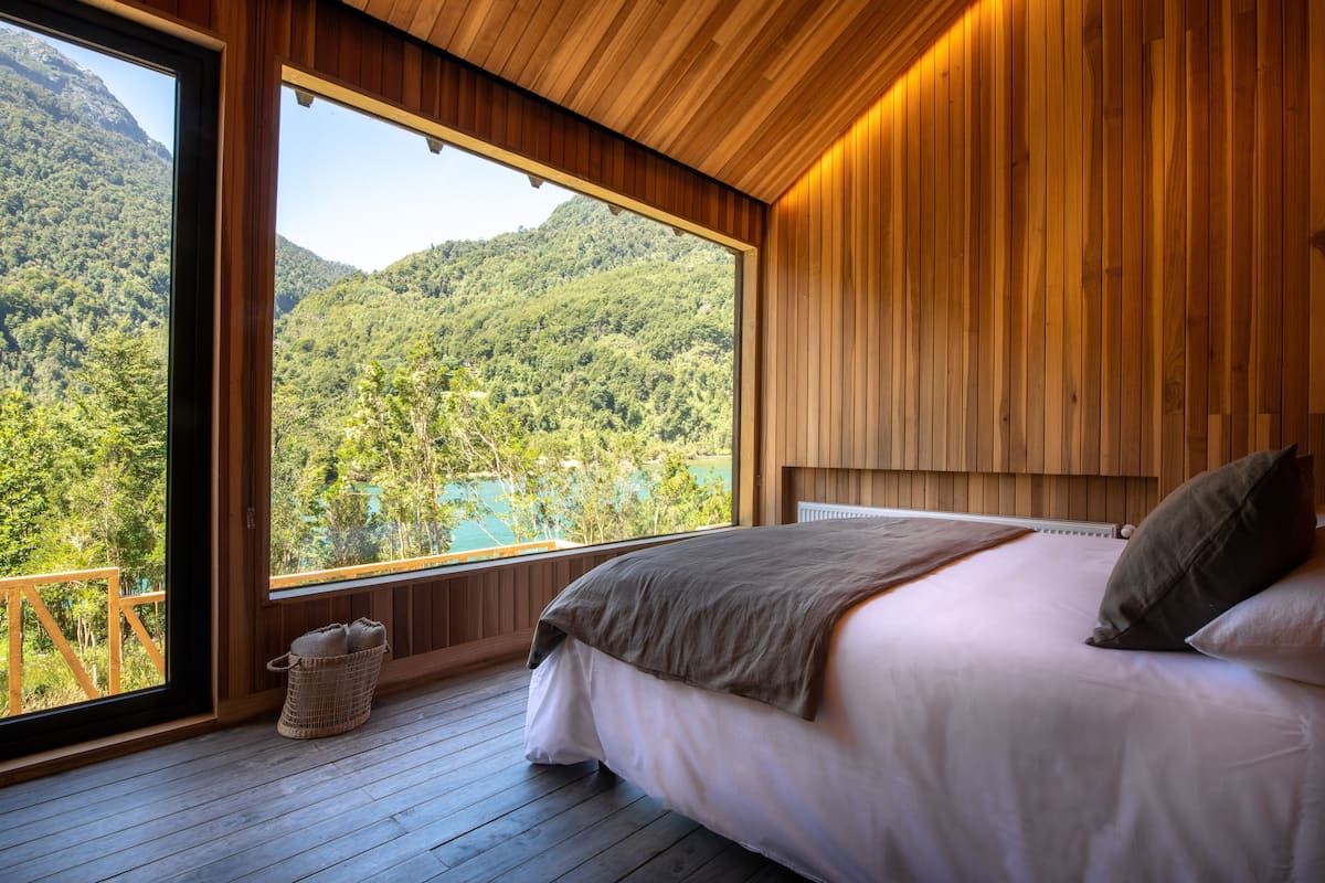 A bedroom with a large window overlooking a forested area and a lake.