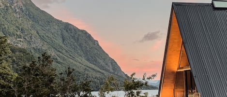 Un edificio moderno con techo inclinado, terraza de madera y vista a un lago y montañas.