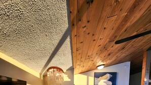 Ein gemütliches Wohnzimmer mit Holzbalkendecke, einem großen Fenster mit Tageslicht, einem modernen Sofa und einem dekorativen Kronleuchter.