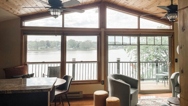 Ein geräumiges Zimmer mit großem Fenster und Seeblick, Deckenventilator, Essbereich mit Tisch und Stühlen sowie eine Sitzecke mit Sofa und Poufs.
