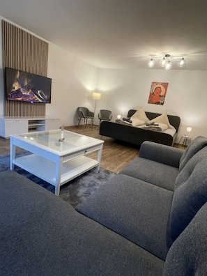 A modern living room with a grey sofa, a white coffee table, and a flat-screen TV mounted on a wooden panel. There is a floor lamp and a painting on the wall.