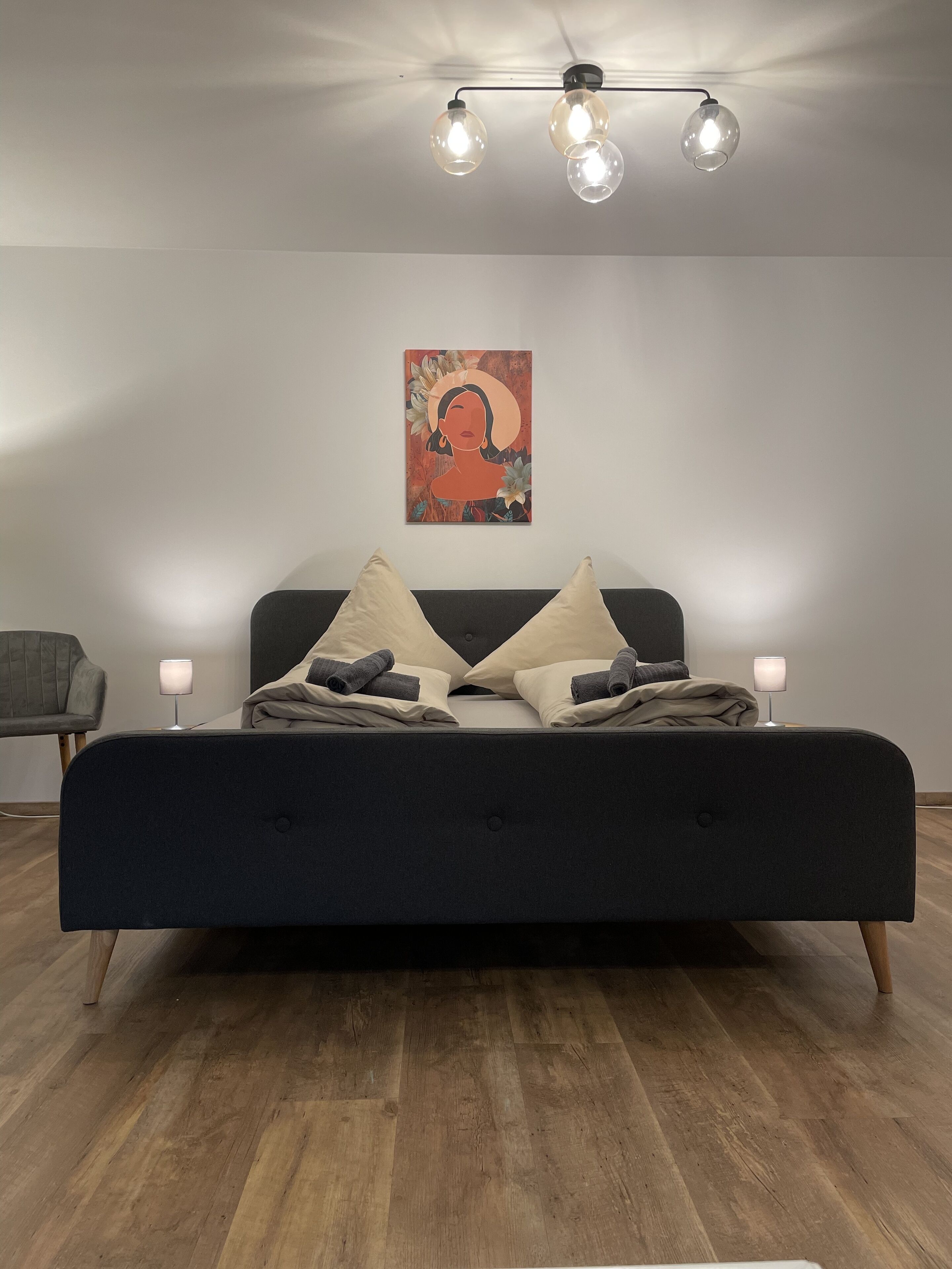 Ett modernt sovrum med en mörk säng, två sänglampor, ett konstverk som hänger på väggen och ett trägolv.