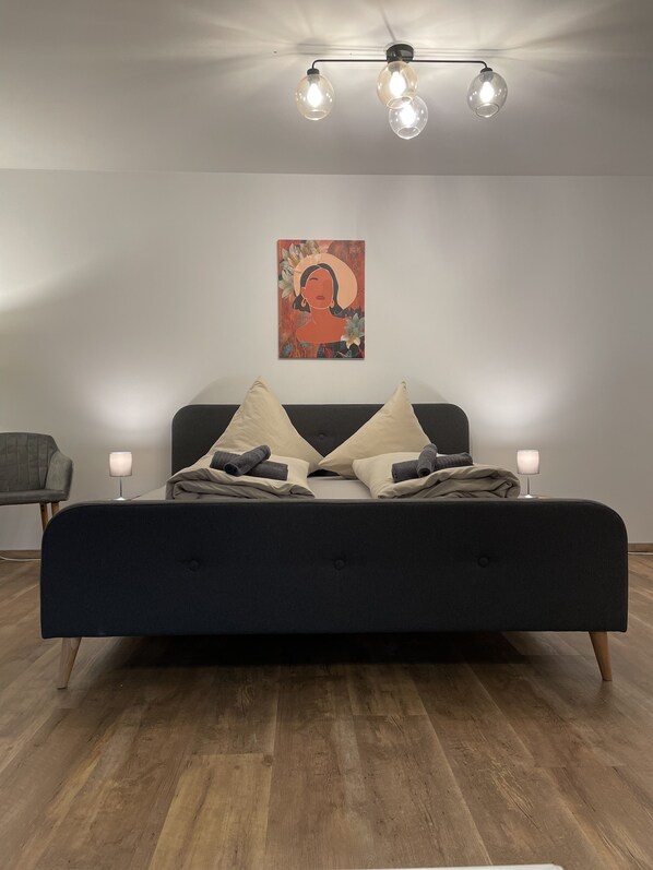 A modern bedroom with a dark-colored bed, two bedside lamps, a wall-mounted artwork, and a wooden floor.