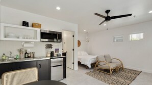 Een kleine keuken met moderne apparatuur, een eethoek met een geweven stoel, een slaapkamer met een bed en een stoel, en een plafondventilator.