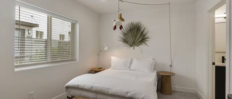 A bedroom with a large bed, a ceiling fan, a bedside table, a nightstand, a hanging plant, and a window with blinds.