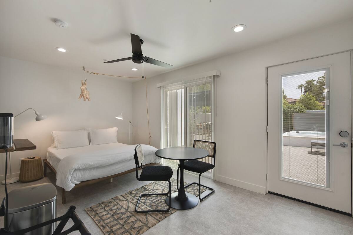 Une chambre à coucher avec un lit, une petite table et des chaises, un ventilateur de plafond et une fenêtre avec des stores.