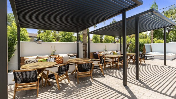 Een terras buiten met houten tafels en stoelen, een bank en een pergola voor beschutting.