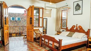 A bedroom with a wooden bed, a chair, a table, and a view of a tiled roof through an open door. - You’ll know that you have rented a historic colonial home when you pass through huge, antique front doors framed by 3 foot-thick adobe walls. Lounge on the shallow, submerged bench seats in the pool or in the hammocks hanging between 12 foot wooden c (Granada)
