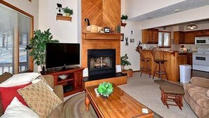 Una sala de estar con chimenea, televisor y una zona de cocina con taburetes de bar.