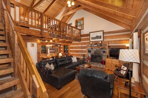 A log cabin interior with a wooden staircase, a fireplace, a black leather sofa, and a dining area.