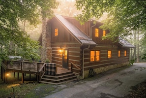 A rustic log cabin with a stone chimney, a wooden deck, and a staircase leading to the entrance.