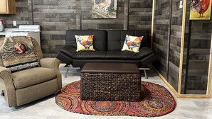 A modern living room with a black leather sofa, two colorful rooster pillows, a woven ottoman, a patterned rug, and a rustic wooden wall. - The Coop.  A lil taste of country with horses and chickens. (Scott)