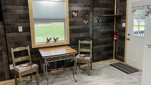 A rustic interior with a wooden wall, a window with a view of a grassy field, two chairs, a small table, and a white door with a fire extinguisher. - The Coop.  A lil taste of country with horses and chickens. (Scott)
