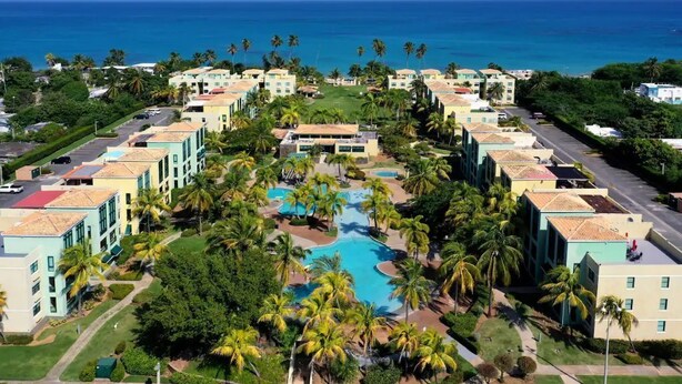 Un resort con varias edificaciones, una piscina central y palmeras.