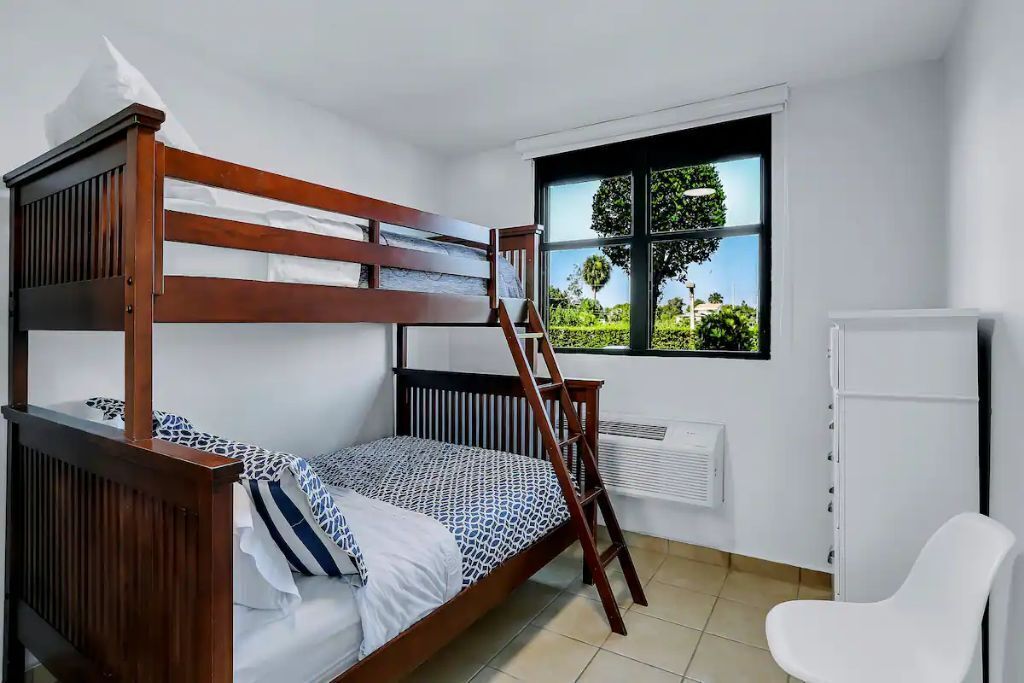 Habitación con literas, una escalera, una silla blanca y una ventana que da a zonas verdes.