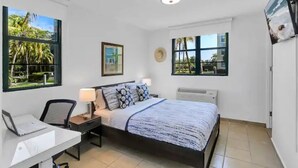 Un dormitorio con cama, escritorio, silla, lámpara y dos ventanas con vistas al verdor.