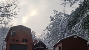 A wooden cabin with a garage and a small shed, surrounded by snow-covered trees.