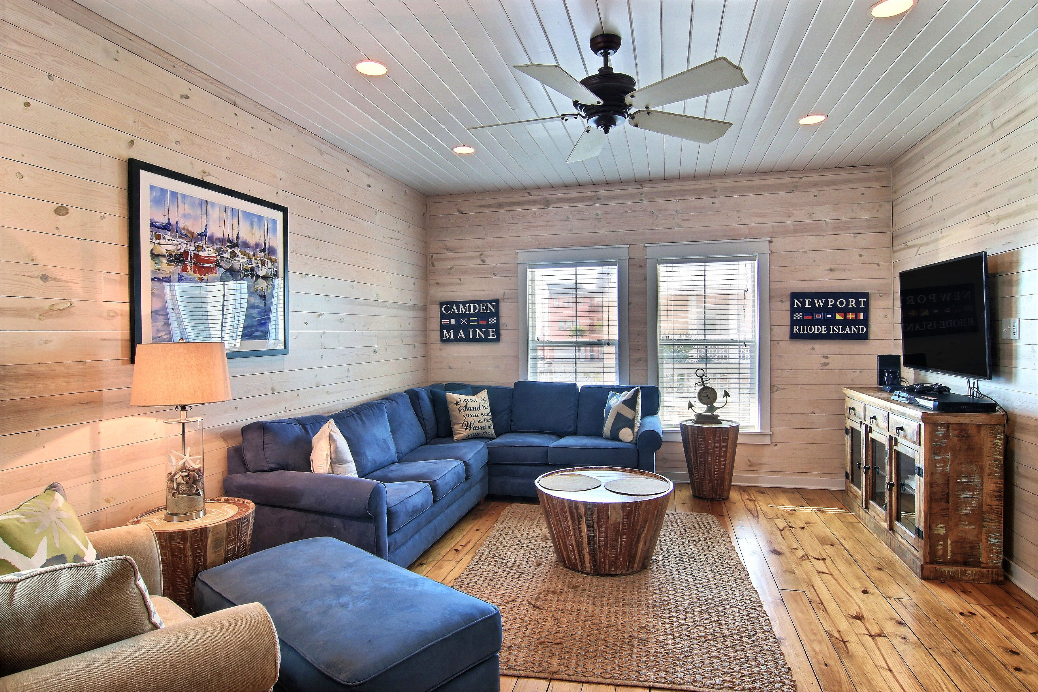 A cozy living room with a blue sectional sofa, a round coffee table, and wooden flooring. There are two windows with blinds, a ceiling fan, and a television on a wooden cabinet.