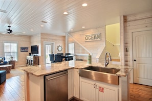Una cocina con encimera de granito, pileta de acero inoxidable y muebles blancos. Hay un cartel en la pared que dice "OCEAN" y una escalera que conduce a otro nivel.