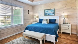 Een slaapkamer met een houten plafond, een groot bed met blauw beddengoed, twee nachtkastjes met lampen en een raam met zonwering.