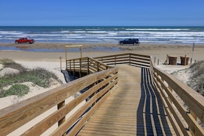 Una pasarela de madera que conduce a una playa, con vehículos estacionados sobre la arena.