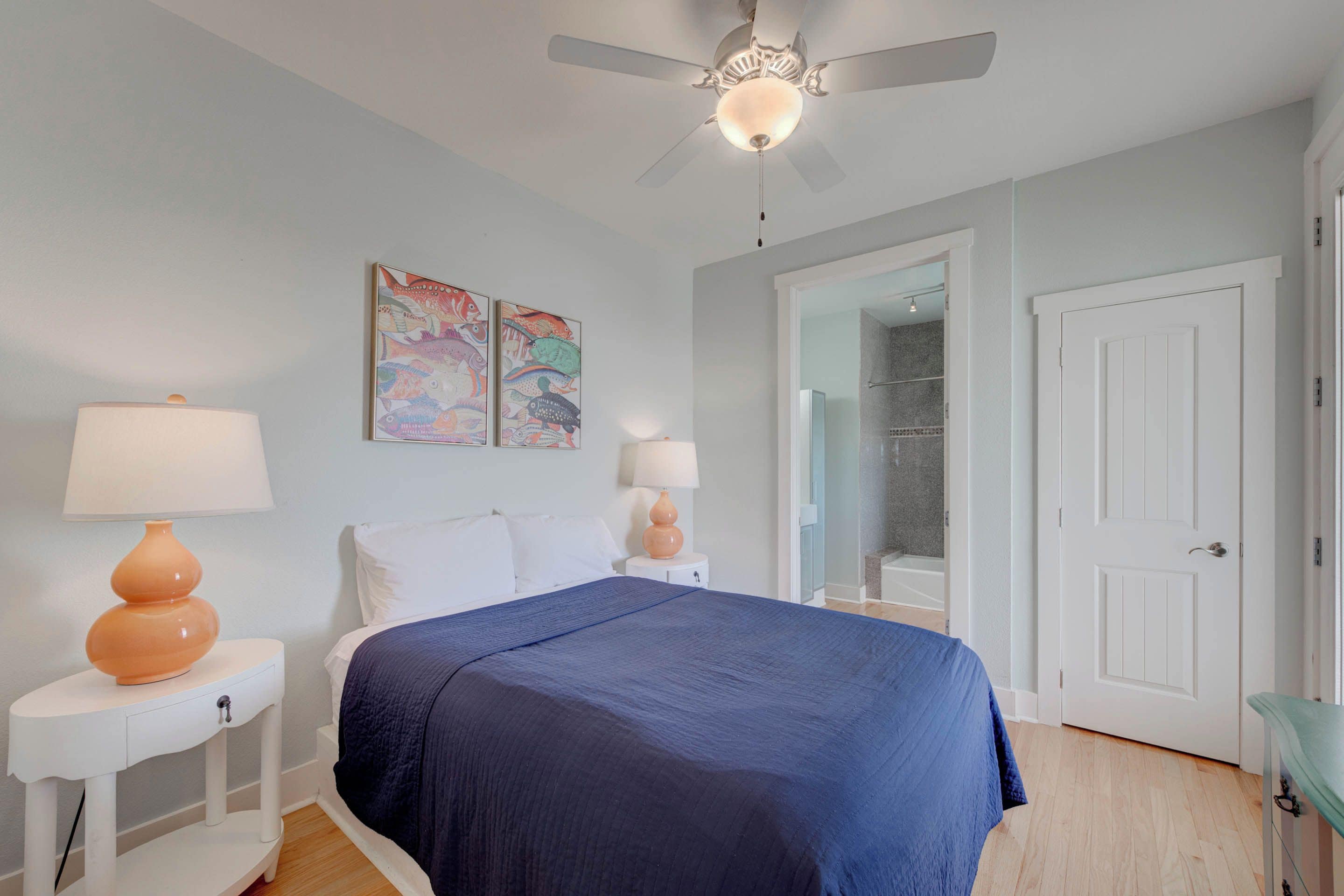 A bedroom with a bed, two bedside tables with lamps, a ceiling fan, and a door to a bathroom.
