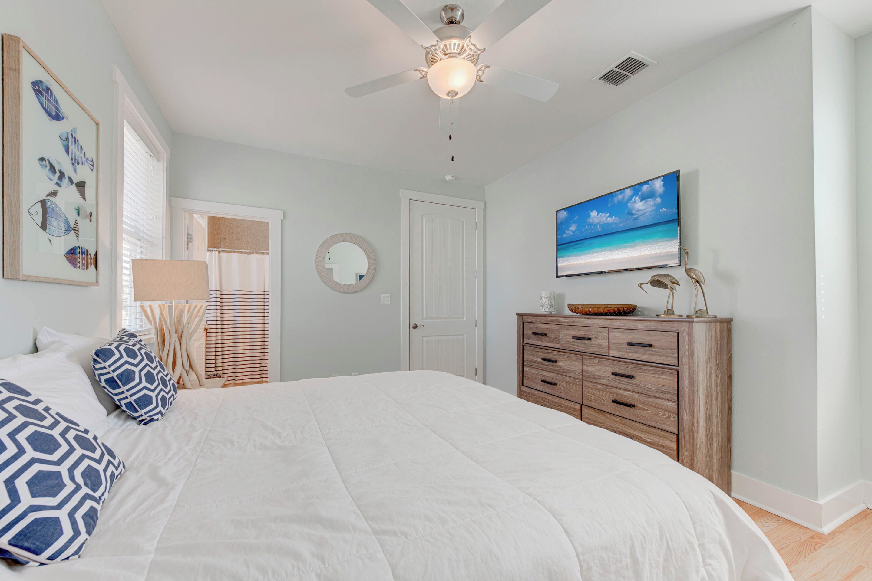A bedroom with a large bed, a ceiling fan, a TV, a wooden dresser, and a framed picture on the wall.