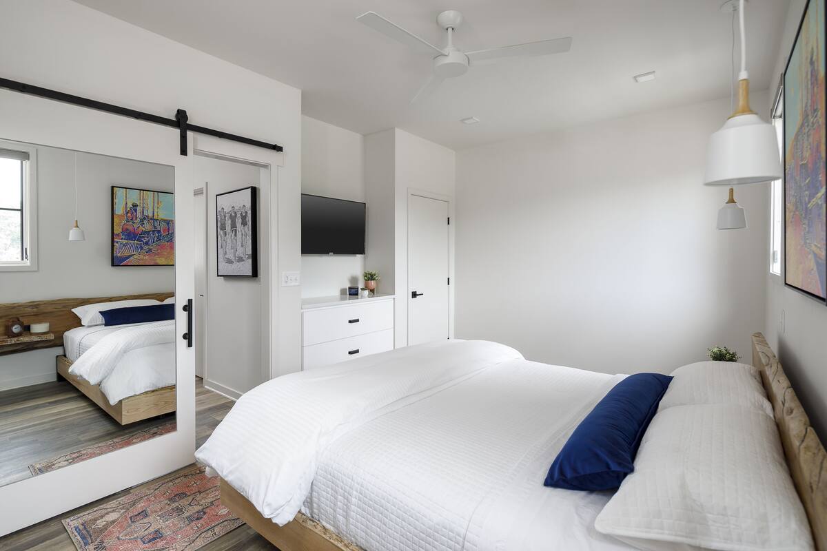 Une chambre à coucher moderne avec un grand lit, un ventilateur de plafond, un téléviseur fixé au mur et une petite plante posée sur la commode.