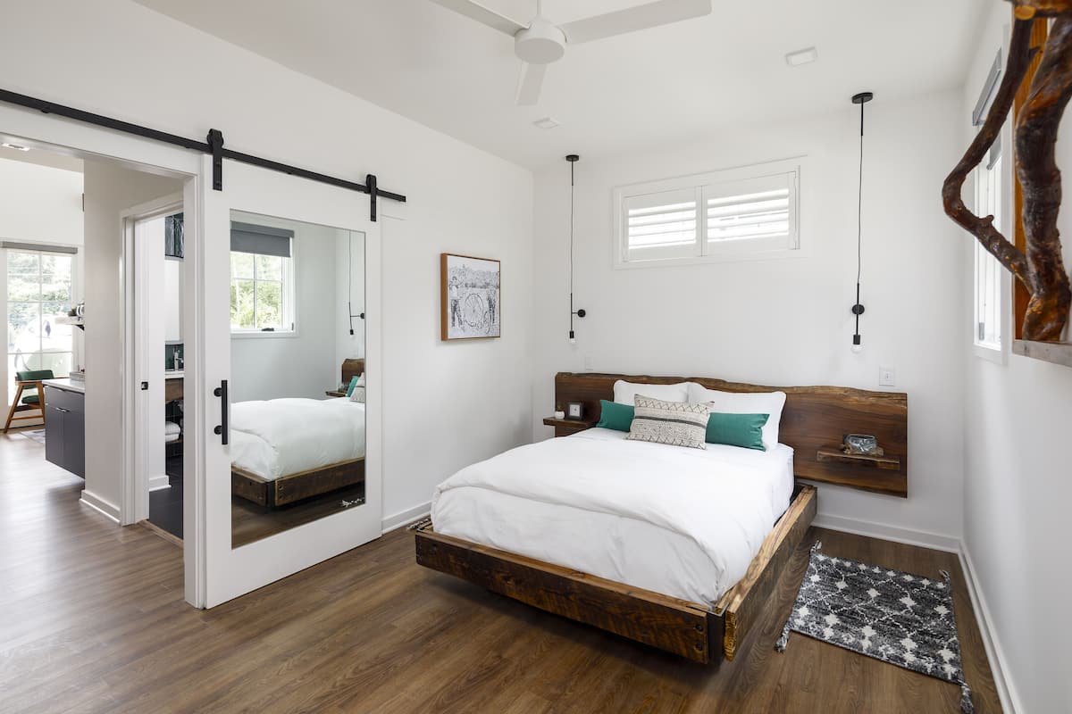 Une chambre avec un lit en bois, une porte coulissante, un ventilateur de plafond, une fenêtre avec des stores et une étagère fixée au mur.