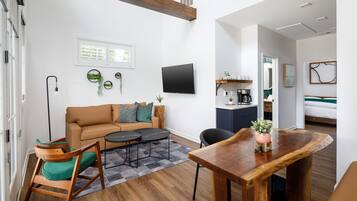 Uma sala de estar moderna com sofá, mesa de centro e mesa de jantar. Há uma TV na parede, uma área de cozinha e um quarto visível ao fundo.