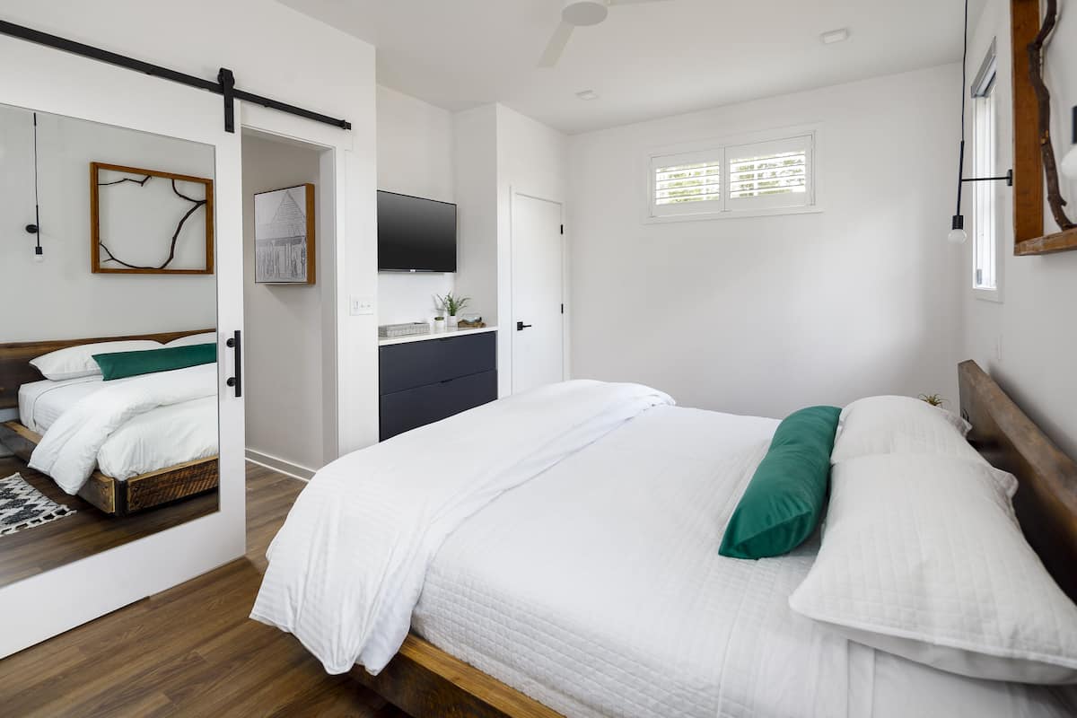 Une chambre à coucher comprenant un lit, une commode, un téléviseur et une fenêtre avec des stores.