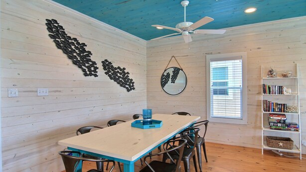 A dining area with a white table and blue chairs, a ceiling fan, and a bookshelf with books and decorative items. - Jumpin' Jellyfish by Silver Sands Vacation Rentals (Port Aransas)